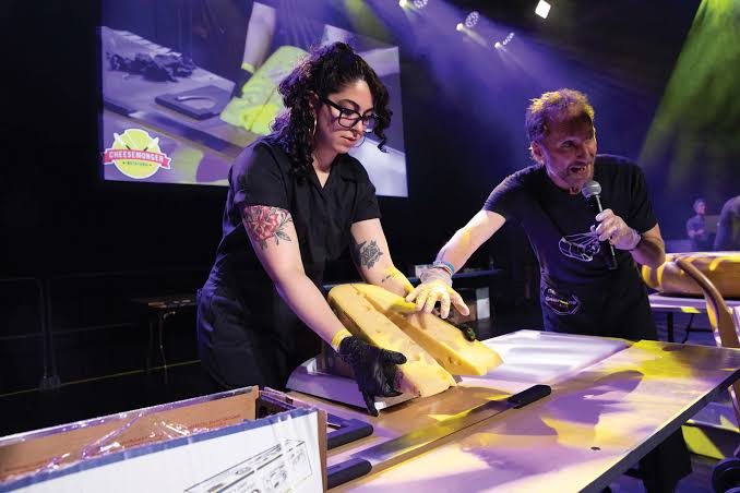 Emilia cutting a wheel of cheese on stage under spotlights