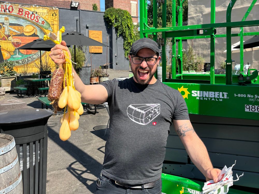 Tommy laughing and holding up strings of caciocavallo and salami in front of the Di Bruno Bros. mural